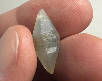 May include: A small, translucent, double-terminated crystal with a diamond-like shape. The stone features a pale, greyish-white hue with subtle internal facets and natural striations along its elongated, pointed structure.