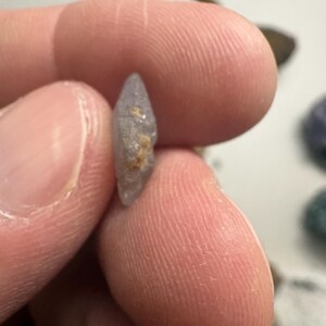 May include: A small, light purple gemstone with a pointed tip and a rough, textured surface. The gemstone is held between two fingers, showcasing its natural shape and color. The stone has a small patch of tan-colored material.