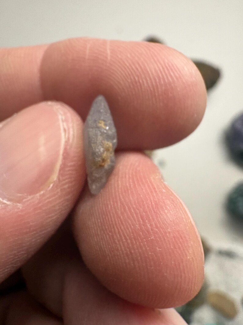 May include: A small, light purple gemstone with a pointed tip and a rough, textured surface. The gemstone is held between two fingers, showcasing its natural shape and color. The stone has a small patch of tan-colored material.