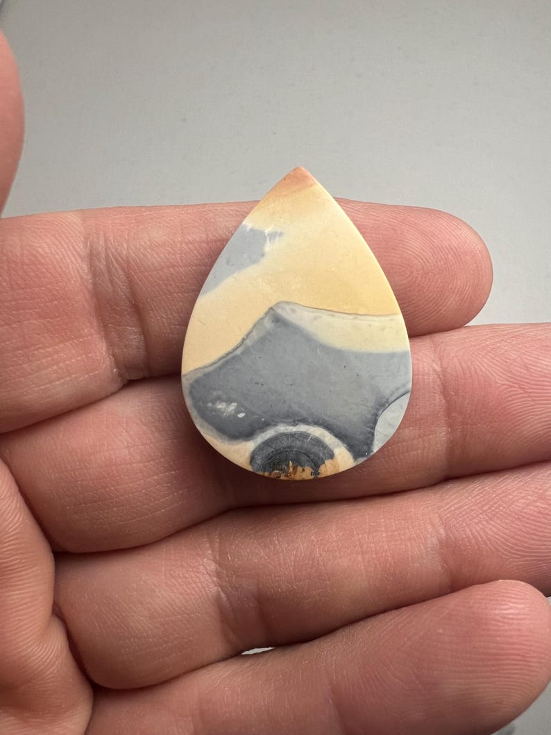 May include: A teardrop-shaped cabochon of polished, multi-colored agate. The stone has a swirling pattern of yellow, gray, and white.