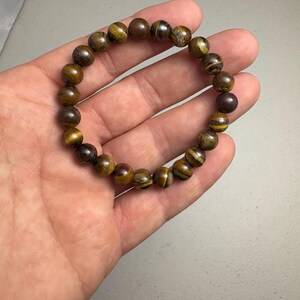 May include: A beaded bracelet featuring round, polished tiger's eye stones in shades of brown and gold. The bracelet is held in a hand, showcasing the natural variations and patterns of the gemstones.
