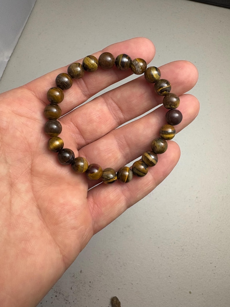 May include: A beaded bracelet featuring round, polished tiger's eye stones in shades of brown and gold. The bracelet is held in a hand, showcasing the natural variations and patterns of the gemstones.