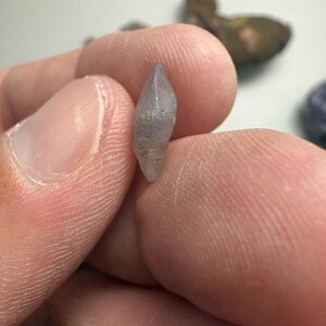 May include: A small, light purple crystal with a pointed tip is held between two fingers. The crystal has a rough, textured surface and appears to be a natural gemstone. Other similar stones are visible in the background.