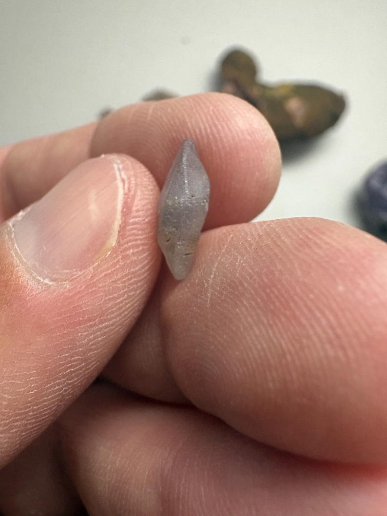 May include: A small, light purple crystal with a pointed tip is held between two fingers. The crystal has a rough, textured surface and appears to be a natural gemstone. Other similar stones are visible in the background.