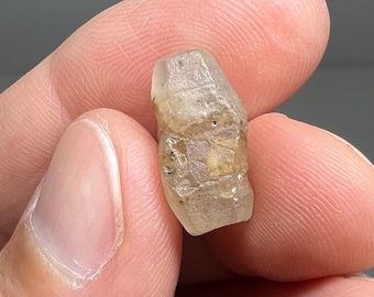 May include: A clear, faceted, light brown gemstone with a rough, natural finish. The gemstone is held in a hand.