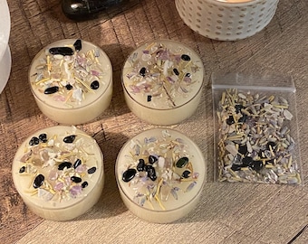 May include: A lit candle in a white ceramic holder with a dotted pattern, surrounded by four small candles with decorative toppings, a black stone, and a small bag of dried herbs and crystals. The candles are on a wooden surface.
