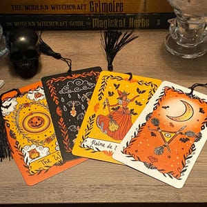 Puede incluir: Cuatro marcap&aacute;ginas decorativos con borlas negras, con ilustraciones de Halloween y brujas. Los marcap&aacute;ginas tienen fondos naranjas y negros con im&aacute;genes de una bruja, murci&eacute;lagos, una luna y una calabaza. El texto "The W" y "Reine de ?" son visibles.