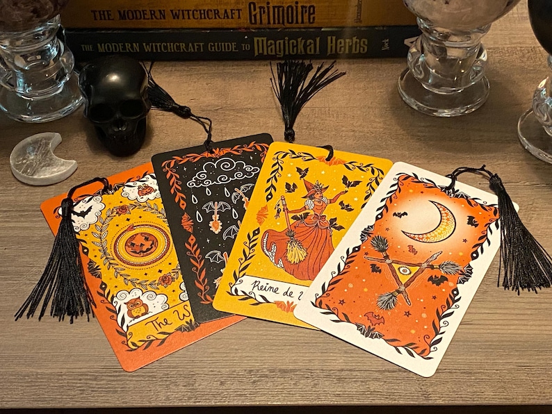 Puede incluir: Cuatro marcap&aacute;ginas decorativos con borlas negras, con ilustraciones de Halloween y brujas. Los marcap&aacute;ginas tienen fondos naranjas y negros con im&aacute;genes de una bruja, murci&eacute;lagos, una luna y una calabaza. El texto "The W" y "Reine de ?" son visibles.
