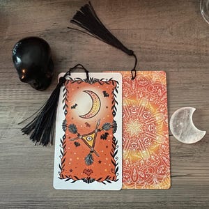Puede incluir: Cartas del tarot de Halloween con un dise&ntilde;o de luna creciente y escoba de bruja, en colores naranja y negro. Tambi&eacute;n hay una calavera negra y un cristal en forma de media luna. Las cartas tienen borlas negras adjuntas.