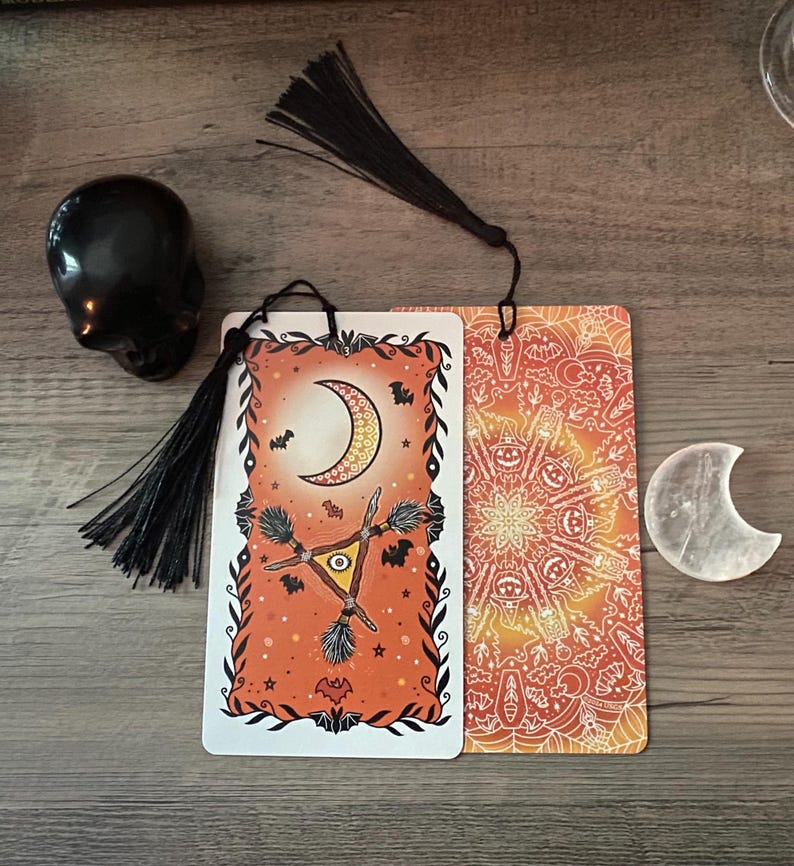 Puede incluir: Cartas del tarot de Halloween con un dise&ntilde;o de luna creciente y escoba de bruja, en colores naranja y negro. Tambi&eacute;n hay una calavera negra y un cristal en forma de media luna. Las cartas tienen borlas negras adjuntas.