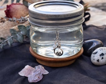 May include: A clear glass jar with a silver metal screw-top lid sits on a circular wooden base. A small silver charm featuring a wave design and a tiny white bead hangs from a silver jump ring attached to the rim of the glass jar.
