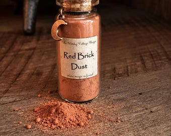 Puede incluir: Una botella de vidrio transparente con tapón de corcho que contiene polvo de ladrillo rojo. Un pequeño dije de media luna color bronce cuelga del cuello. La etiqueta dice: The Witchy Cottage Shoppe, Red Brick Dust, where magic is found. Un montón de polvo rojo se encuentra delante.