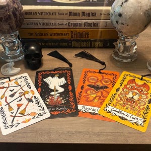 Puede incluir: Un conjunto de marcap&aacute;ginas decorativos con borlas negras, con ilustraciones de escobas, murci&eacute;lagos y dise&ntilde;os de cartas del tarot. Los marcap&aacute;ginas est&aacute;n colocados sobre una superficie de madera, junto con esferas de cristal, libros y una calavera negra.