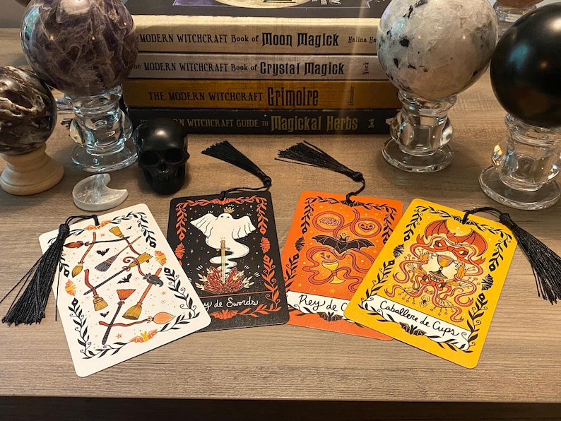 Puede incluir: Un conjunto de marcap&aacute;ginas decorativos con borlas negras, con ilustraciones de escobas, murci&eacute;lagos y dise&ntilde;os de cartas del tarot. Los marcap&aacute;ginas est&aacute;n colocados sobre una superficie de madera, junto con esferas de cristal, libros y una calavera negra.