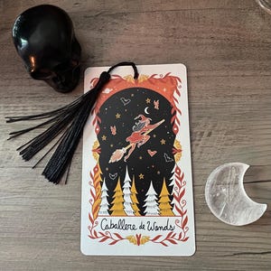 Puede incluir: Una calavera negra, una tarjeta decorativa con una bruja en una escoba y un cristal en forma de media luna se exhiben sobre una superficie de madera. La tarjeta presenta el texto "Caballere de Wands" y una borla negra.