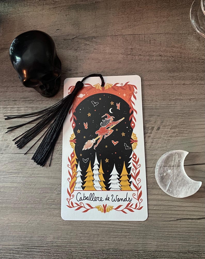 Puede incluir: Una calavera negra, una tarjeta decorativa con una bruja en una escoba y un cristal en forma de media luna se exhiben sobre una superficie de madera. La tarjeta presenta el texto "Caballere de Wands" y una borla negra.