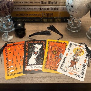 Puede incluir: Cuatro marcap&aacute;ginas de cartas del tarot con borlas negras. Las cartas presentan ilustraciones con dise&ntilde;os en naranja, negro y blanco. Una carta dice "The Lovers", otra "Page of Pentacles" y una tercera "Ace of Wands".