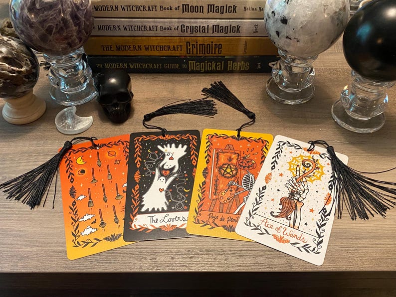 Puede incluir: Cuatro marcap&aacute;ginas de cartas del tarot con borlas negras. Las cartas presentan ilustraciones con dise&ntilde;os en naranja, negro y blanco. Una carta dice "The Lovers", otra "Page of Pentacles" y una tercera "Ace of Wands".