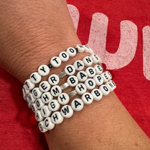 May include: A stack of white beaded bracelets with black lettering spelling out the words "Too High", "Danger", "Babe", "Hope", and "Schwarbomb".
