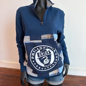 May include: A navy blue long-sleeve pullover with a quarter-zip neckline. The front features a patchwork design with a large circular emblem that reads "PHILADELPHIA UNION" and a snake logo. The garment is displayed on a mannequin.