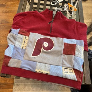 May include: A red quarter-zip sweatshirt with a gray and patchwork panel featuring the Philadelphia Phillies logo. The patchwork includes various fabrics in shades of blue, red, and gray. The sweatshirt is folded on a wooden table.