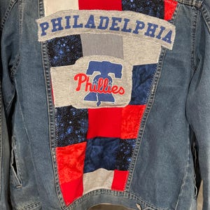 May include: A denim jacket with a patchwork design on the back. The patches include red, white, blue, and gray fabrics. The word "PHILADELPHIA" is across the top, and the Phillies logo is in the center.