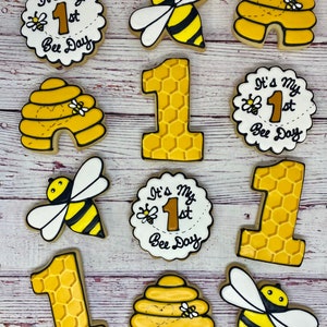 May include: A collection of decorated sugar cookies with a bee theme. The cookies include bee-shaped designs, honeycomb patterns, and cookies with the text "It's my 1st Bee Day". The cookies are yellow, black, white, and brown.