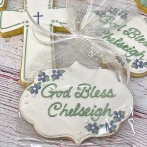 May include: A white cookie with green and purple icing. The cookie is shaped like a banner and says "God Bless Chelseigh".