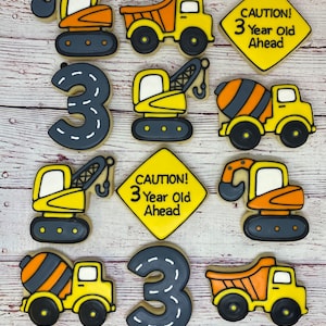 May include: A set of ten decorated sugar cookies featuring construction vehicles and the number three. The cookies are yellow, orange, black, and gray. The cookies include a dump truck, a cement mixer, an excavator, and a sign that reads "CAUTION! 3 Year Old Ahead".