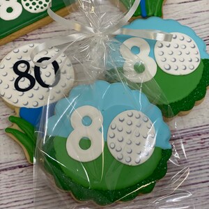 May include: A set of four golf-themed cookies decorated with green, blue and white icing. The cookies feature the number 80 and golf balls. The cookies are packaged in a clear plastic bag tied with a white ribbon.