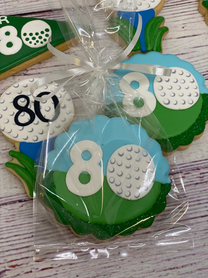 May include: A set of four golf-themed cookies decorated with green, blue and white icing. The cookies feature the number 80 and golf balls. The cookies are packaged in a clear plastic bag tied with a white ribbon.