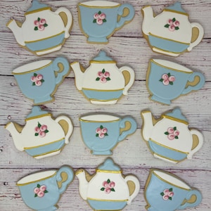 May include: A collection of decorated sugar cookies in the shape of teacups and teapots. The cookies are light blue and white with gold trim and pink floral accents. The cookies are arranged on a light wood-grain surface.