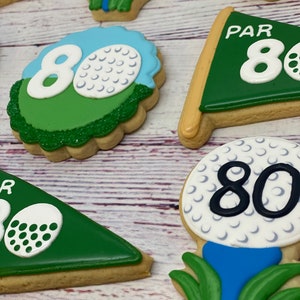 May include: A set of five golf-themed sugar cookies decorated with green, blue, white, and black icing. The cookies feature golf balls, golf flags, and the number 80.