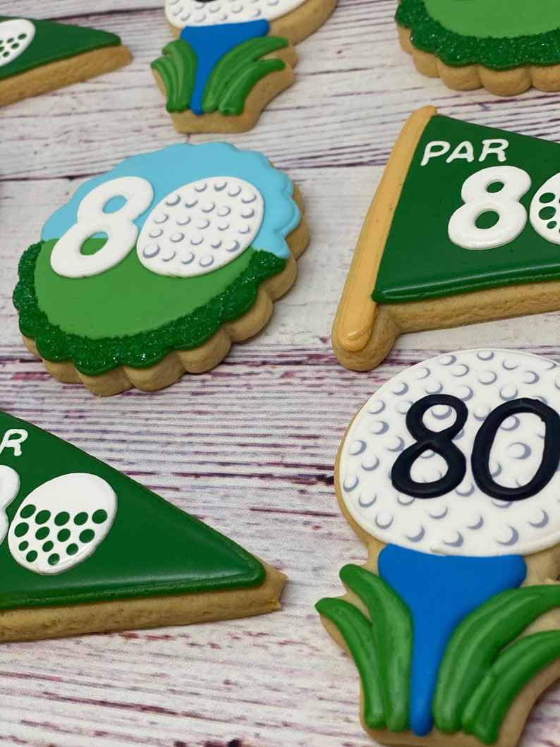 May include: A set of five golf-themed sugar cookies decorated with green, blue, white, and black icing. The cookies feature golf balls, golf flags, and the number 80.