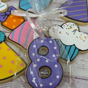May include: A purple number eight cookie with white polka dots, decorated with black icing, packaged in a clear plastic bag with a white ribbon.