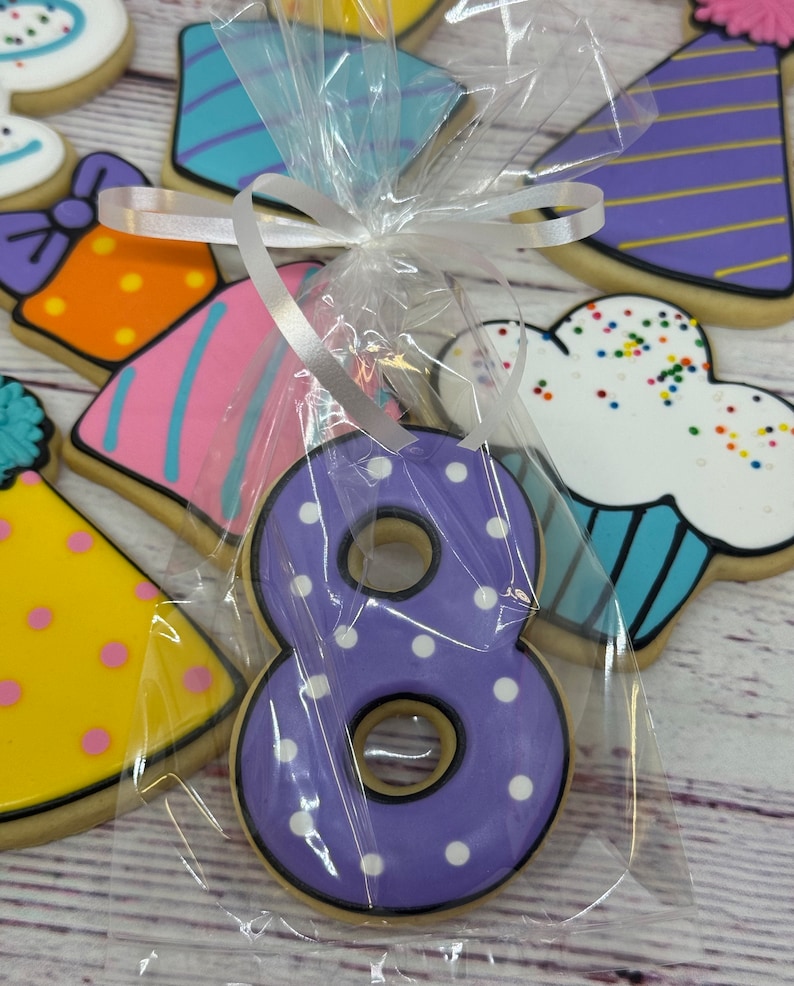 May include: A purple number eight cookie with white polka dots, decorated with black icing, packaged in a clear plastic bag with a white ribbon.
