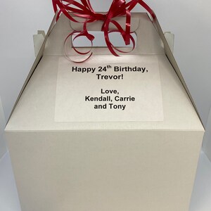 May include: A white gift box with a red ribbon on top. The box has a label that says "Happy 24th Birthday, Trevor! Love, Kendall, Carrie and Tony."