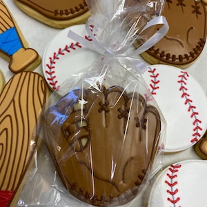 Puede incluir: Una galleta de guante de b&eacute;isbol marr&oacute;n decorada con costuras blancas y una cinta blanca atada alrededor de una bolsa de pl&aacute;stico transparente. La galleta est&aacute; rodeada de otras galletas con tem&aacute;tica de b&eacute;isbol, incluyendo una pelota de b&eacute;isbol, un bate y otro guante.