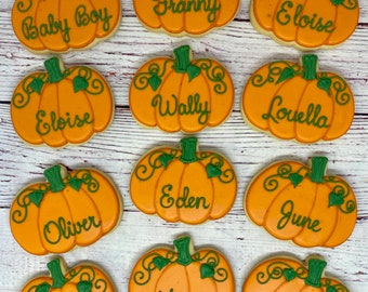May include: Twelve orange pumpkin-shaped cookies decorated with green vines and leaves. Each cookie is decorated with a different name, including "Baby Boy", "Franny", "Eloise", "Wally", "Louella", "Eden", "June", "Oliver", "Rose", "Maeve", and "Vivian".