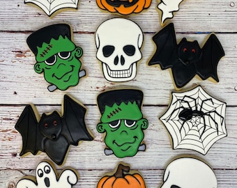 May include: A collection of 12 Halloween-themed sugar cookies decorated with various designs, including pumpkins, ghosts, bats, spiders, and skulls. The cookies are arranged in three rows on a white wooden background.