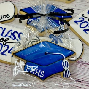 May include: A blue graduation cap cookie with white icing and the text "FMHS" on it. The cookie is packaged in a clear plastic bag with a black ribbon.