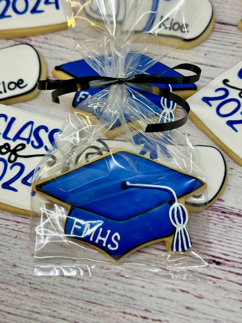 May include: A blue graduation cap cookie with white icing and the text "FMHS" on it. The cookie is packaged in a clear plastic bag with a black ribbon.