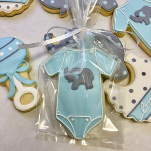May include: A blue baby onesie cookie decorated with a grey elephant, packaged in a clear plastic bag with a white ribbon. The cookie is surrounded by other baby-themed cookies, including a rattle, a bib, and an elephant.