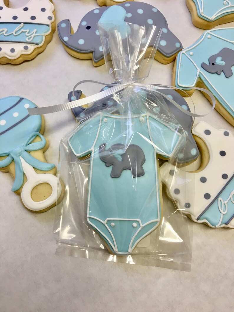 May include: A blue baby onesie cookie decorated with a grey elephant, packaged in a clear plastic bag with a white ribbon. The cookie is surrounded by other baby-themed cookies, including a rattle, a bib, and an elephant.