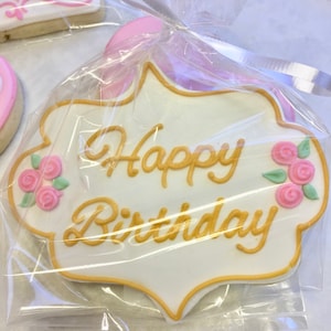 以下が含まれることがあります： 白いクッキーに、金色のアイシングで「Happy Birthday」と書かれています。クッキーには、ピンクのバラと緑の葉も飾られています。クッキーは透明なプラスチックで包まれています。