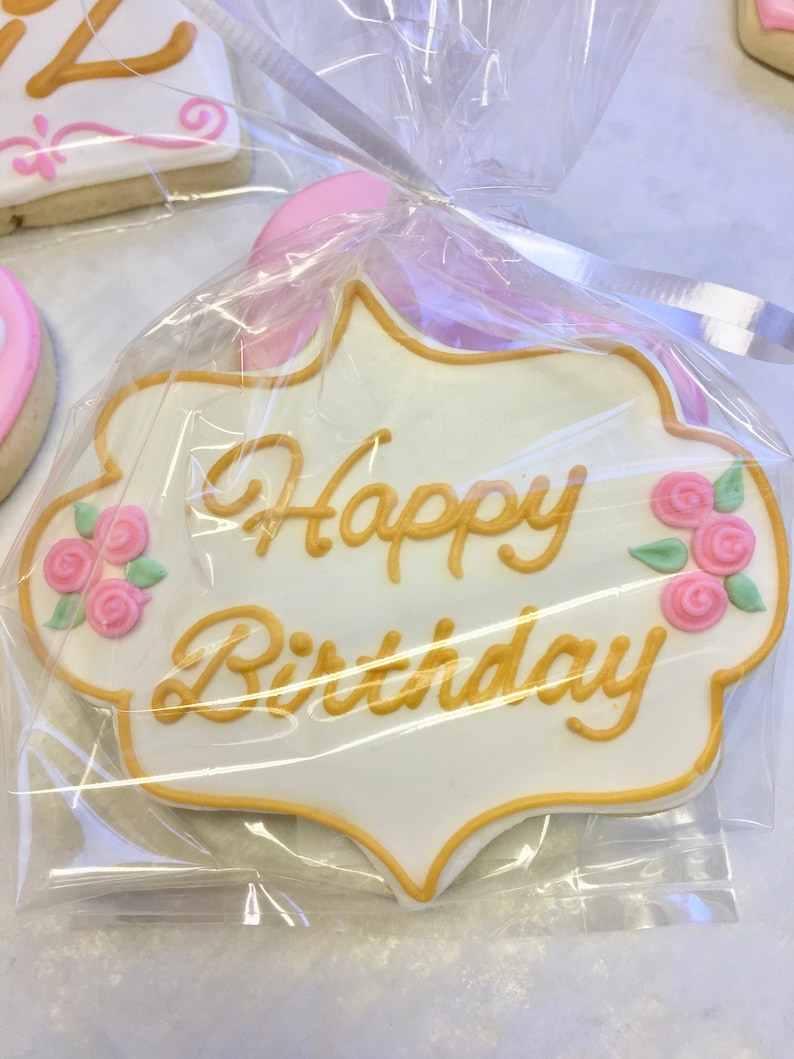 以下が含まれることがあります： 白いクッキーに、金色のアイシングで「Happy Birthday」と書かれています。クッキーには、ピンクのバラと緑の葉も飾られています。クッキーは透明なプラスチックで包まれています。