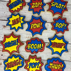 May include: Twelve decorated sugar cookies with various comic book sound effects, such as "WHAM!" and "POW!" in red, yellow, and blue.