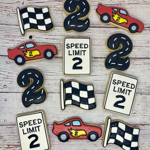May include: Nine decorated sugar cookies with a racing theme. The cookies are decorated with red race cars, black and white checkered flags, black number twos, and speed limit signs that say "SPEED LIMIT 2".
