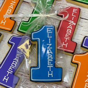 May include: A blue and white decorated sugar cookie in the shape of the number one with the name "Elizabeth" written vertically on it. The cookie is packaged in a clear plastic bag with a white ribbon.