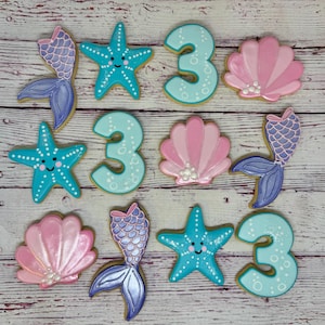 May include: A collection of mermaid-themed sugar cookies. The cookies include blue starfish with smiling faces, light blue number 3 cookies, pink seashell cookies, and mermaid tail cookies in shades of purple and blue.